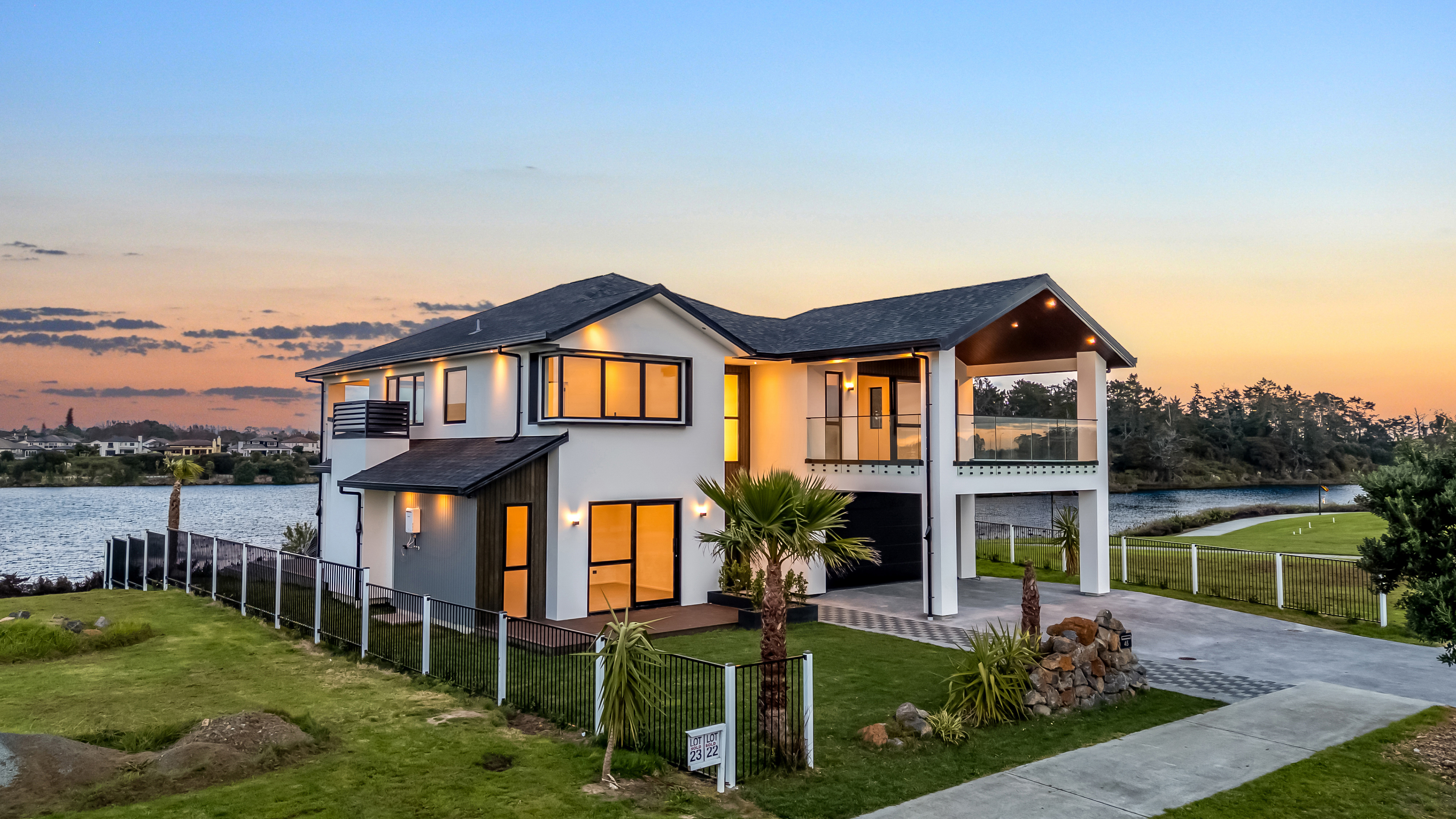45 Severina Drive, Karaka, 4 bedroom House. Modern white two-story design with dark roof, balconies, and waterfront views at sunset.